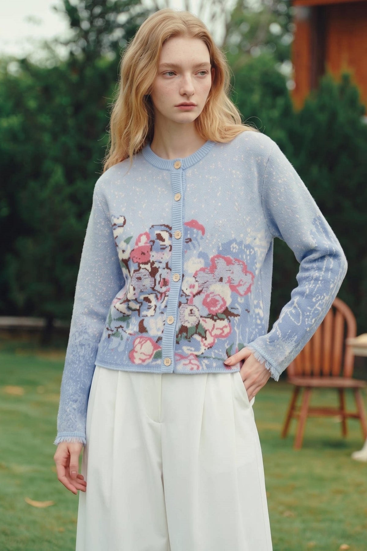 A woman standing outdoors in a garden wearing a light blue knit cardigan with a large floral graphic on the front, white wide-leg pants, and holding one hand in her pocket.
