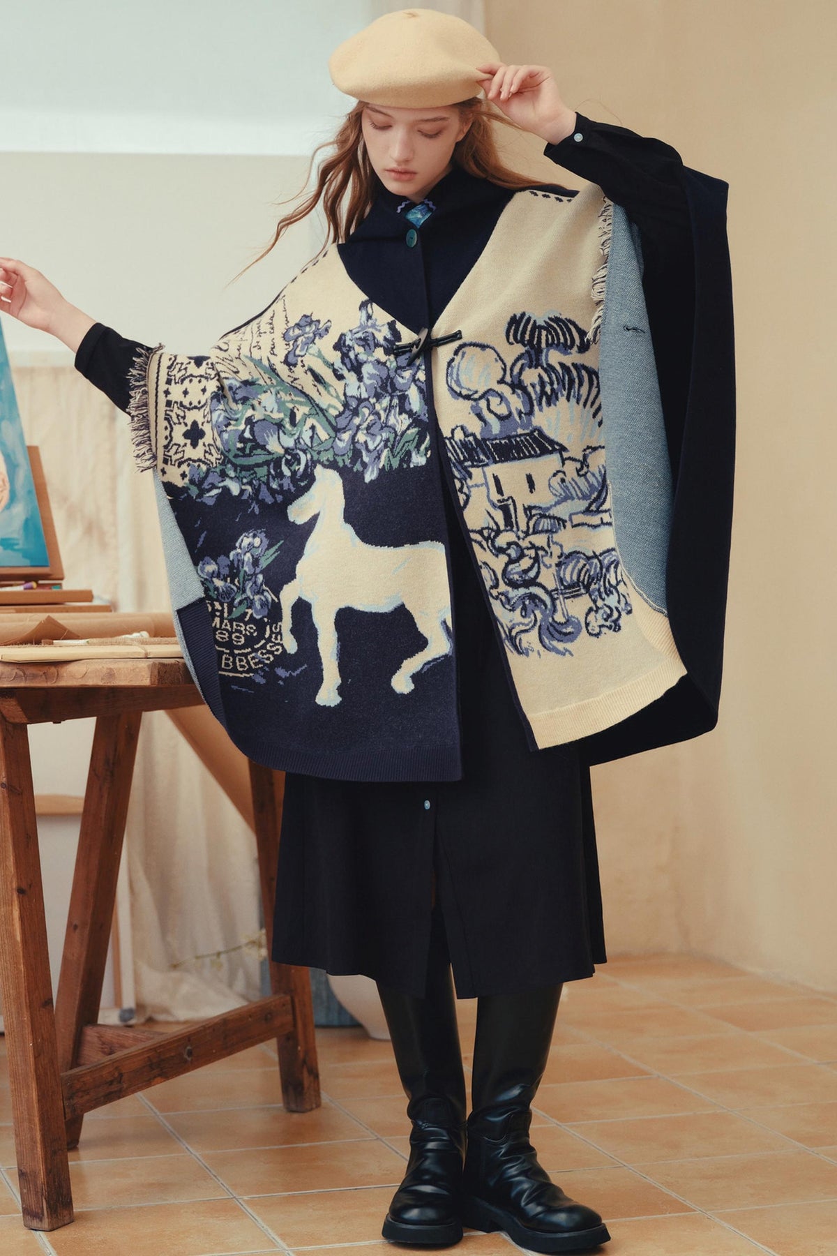 A woman models a large, draped knit poncho with a complex graphic pattern featuring a white dog, blue floral motifs, and a black cat, styled with a black coat, beret, and boots in an indoor setting.