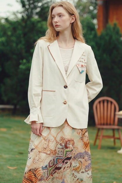 A woman stands outdoors on grass wearing a cream-colored blazer with decorative embroidery and a long, patterned skirt. The background features green foliage and a wooden chair.