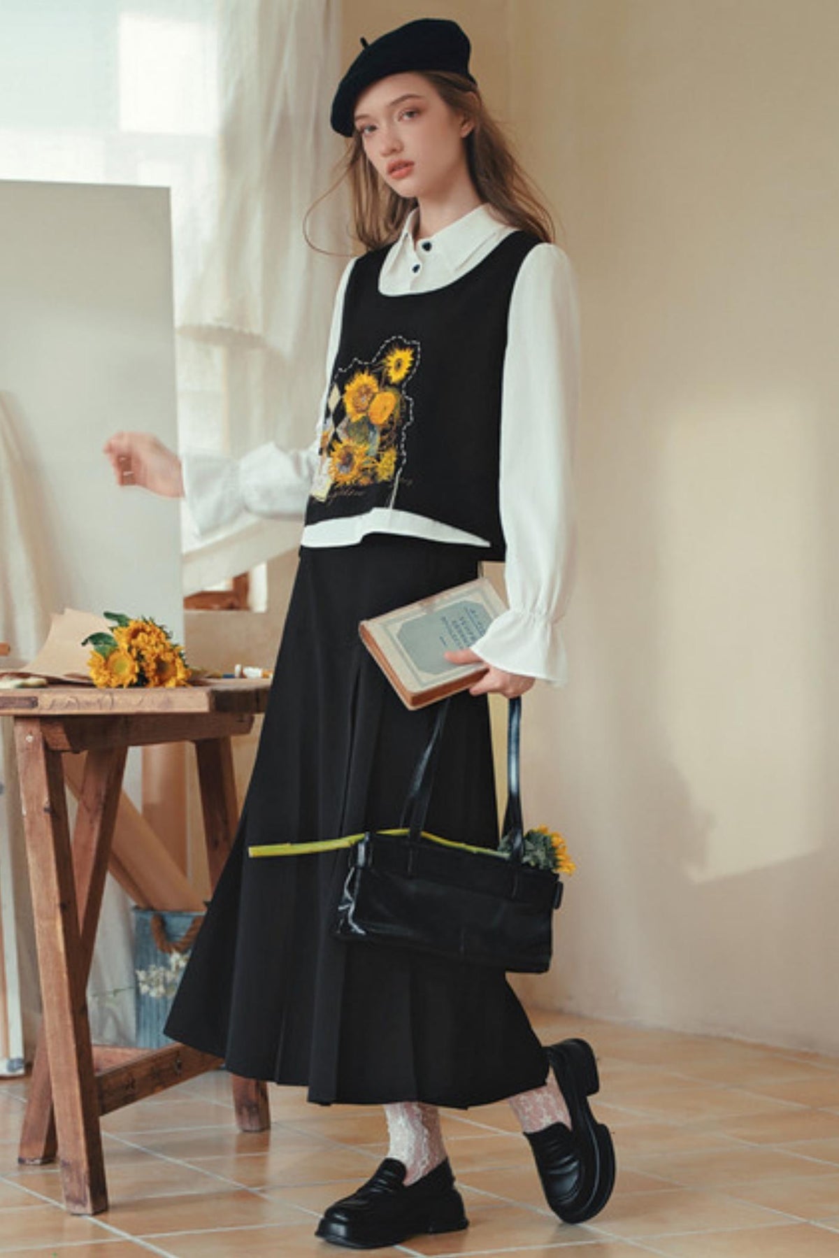 A woman models a black sleeveless vest and black pleated skirt set with a white collared shirt, black beret, black loafers, and a black handbag in a warm, sunlit studio setting.