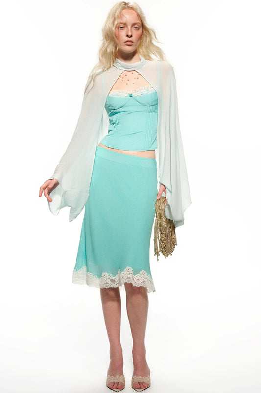 A blonde model wearing a light aqua two-piece set consisting of a lace-trimmed camisole top and matching midi skirt, styled with a sheer white draped capelet and holding a gold fringe clutch against a white studio background.