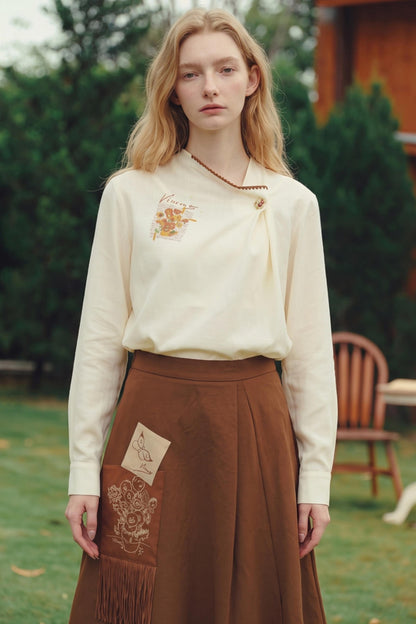 A woman stands outdoors wearing a cream long-sleeve blouse with floral embroidery and a brown pleated skirt with fringe and patchwork details.