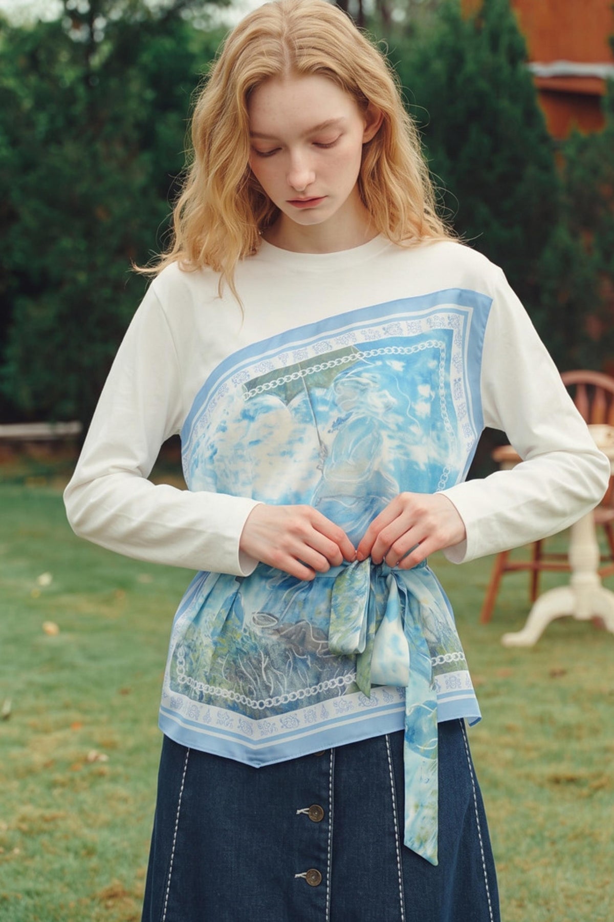 A woman outdoors wearing a white long-sleeve top with a large light blue and white printed panel across the chest, tied at the waist with a matching fabric tie, paired with a dark denim skirt.