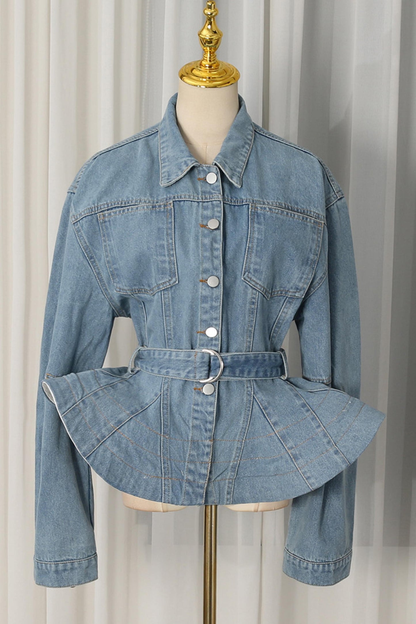 A light blue denim jacket with a unique peplum waist and matching belt, displayed on a cream dress form with a gold finial against a white curtain background.
