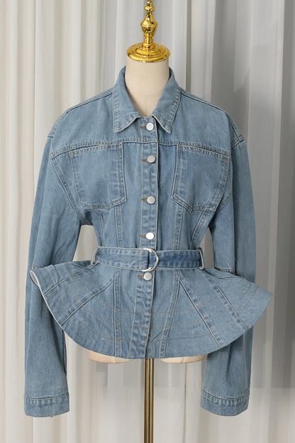 A light blue denim jacket with a unique peplum waist and matching belt, displayed on a cream dress form with a gold finial against a white curtain background.