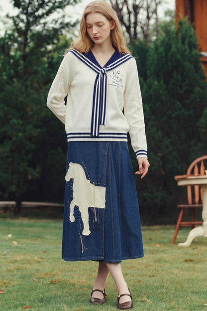 A woman stands outdoors on grass wearing a cream knit cardigan with navy trim and a matching navy midi skirt featuring a large cream horse applique. Trees and a wooden chair are visible in the background.