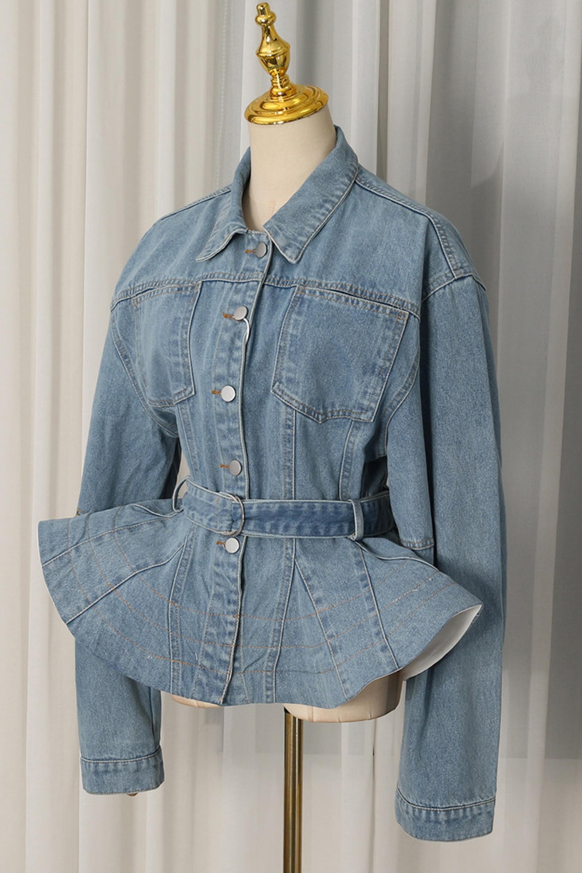A light blue denim jacket with a matching self-tie belt, displayed on a cream dress form with a gold finial against a white curtain backdrop.