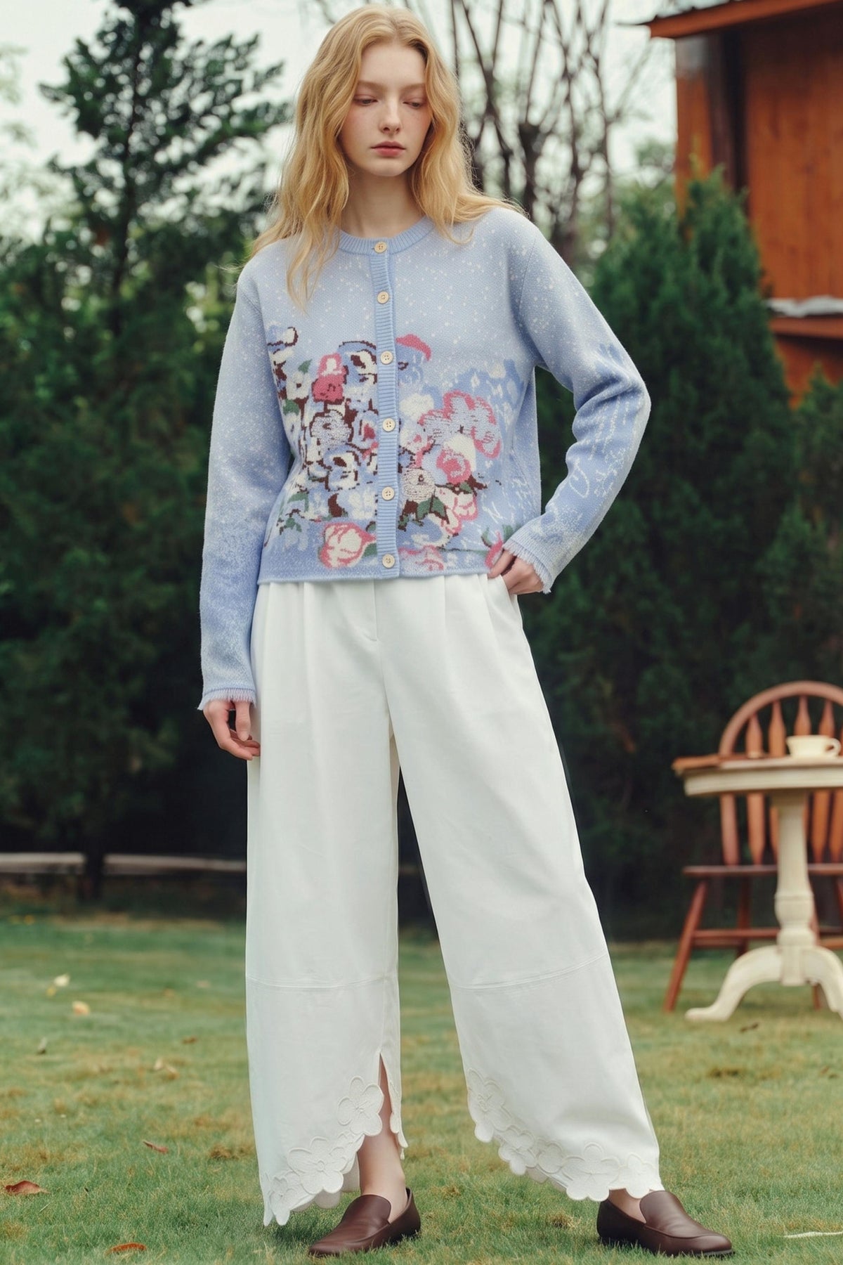 A woman stands outdoors on grass wearing a light blue cardigan with a colorful floral and character print across the front, paired with wide-leg white pants and brown shoes.