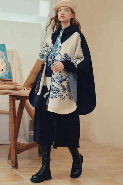 A woman models a stylish outfit featuring a cream and navy blue patterned cape jacket over a black dress, paired with a beige beret and black boots, in a warm-toned indoor setting.