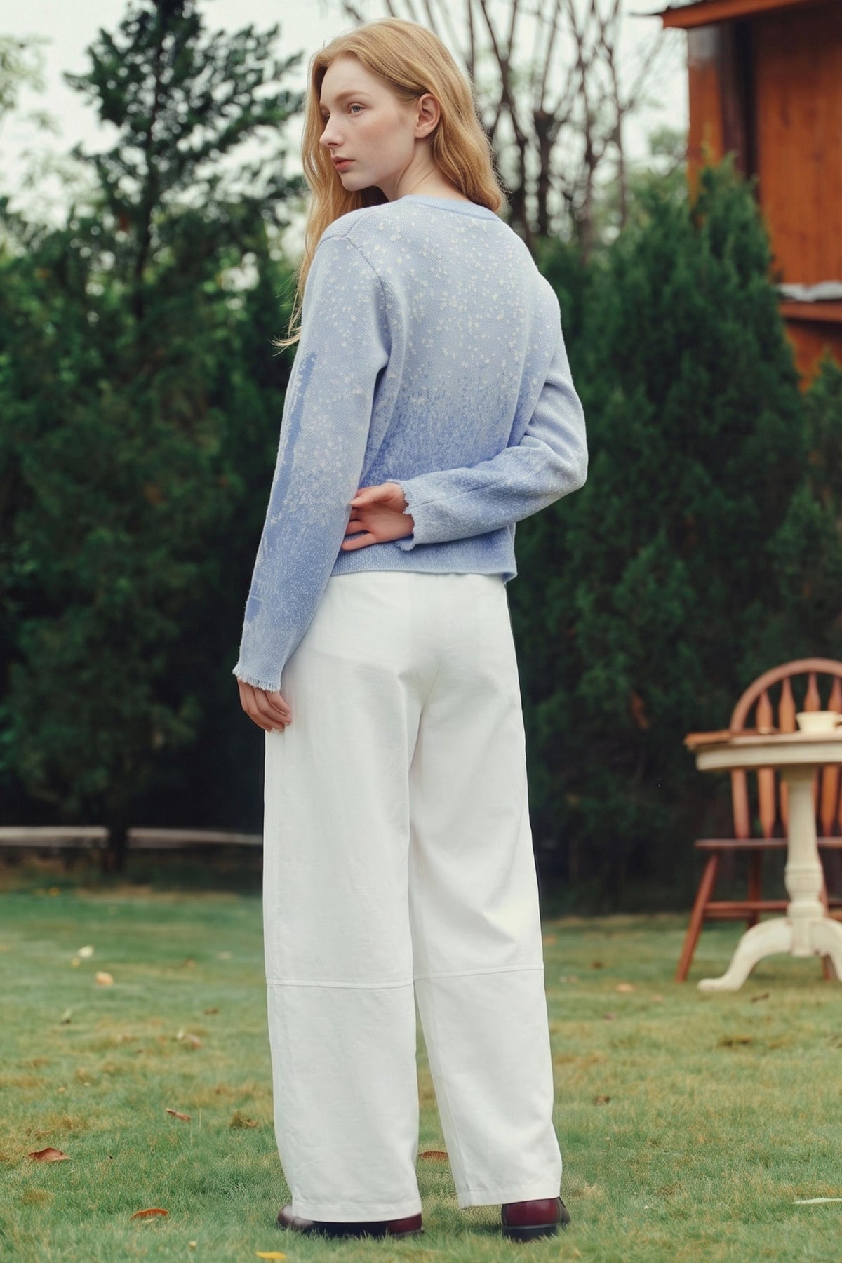 A woman stands outdoors on a grassy lawn wearing a light blue knit sweater and white wide-leg pants, photographed from the back.