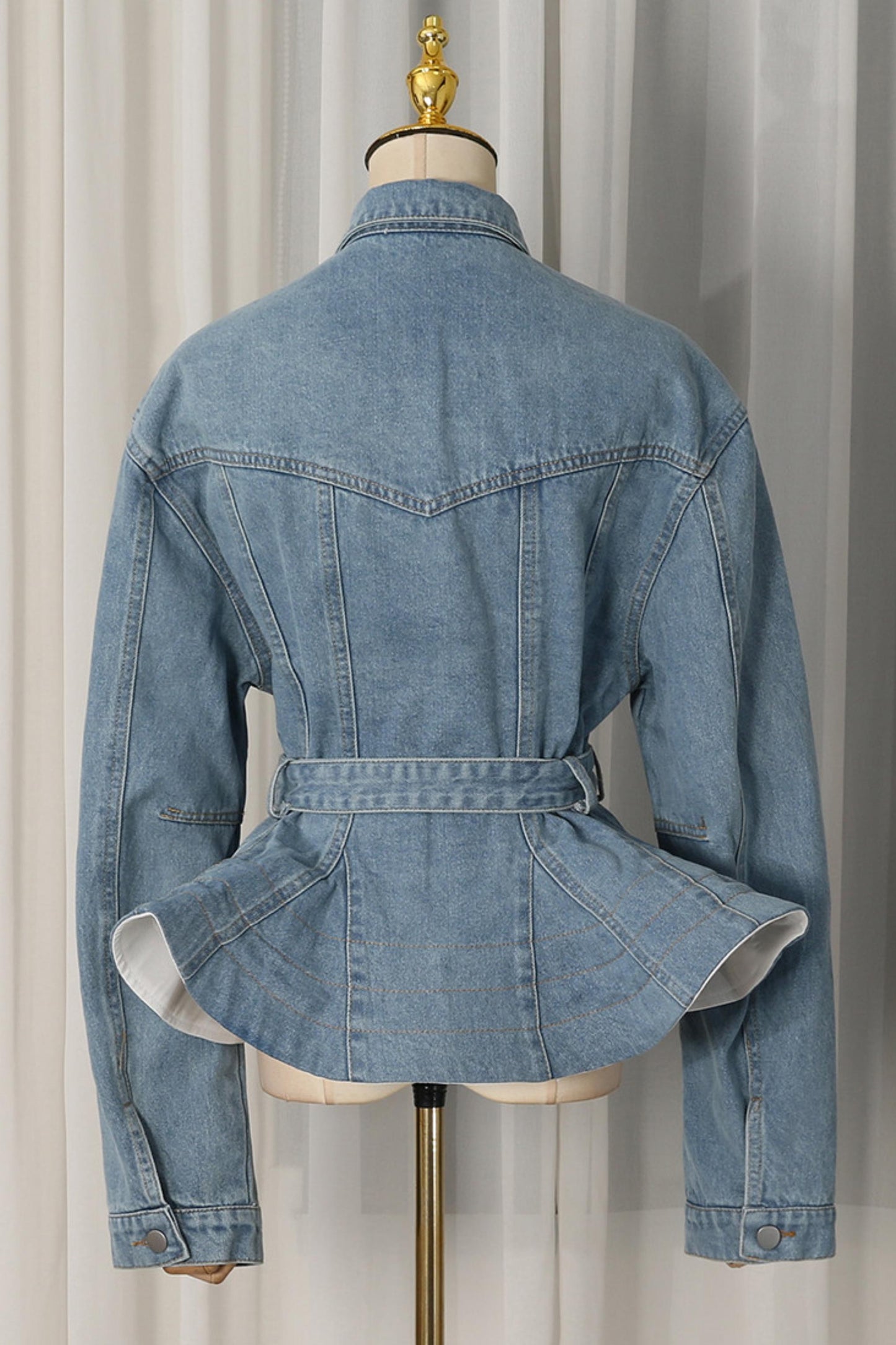 A light blue denim jacket displayed on a dress form mannequin, showing the back view with a peplum hem and waist belt, set against a neutral curtain background.