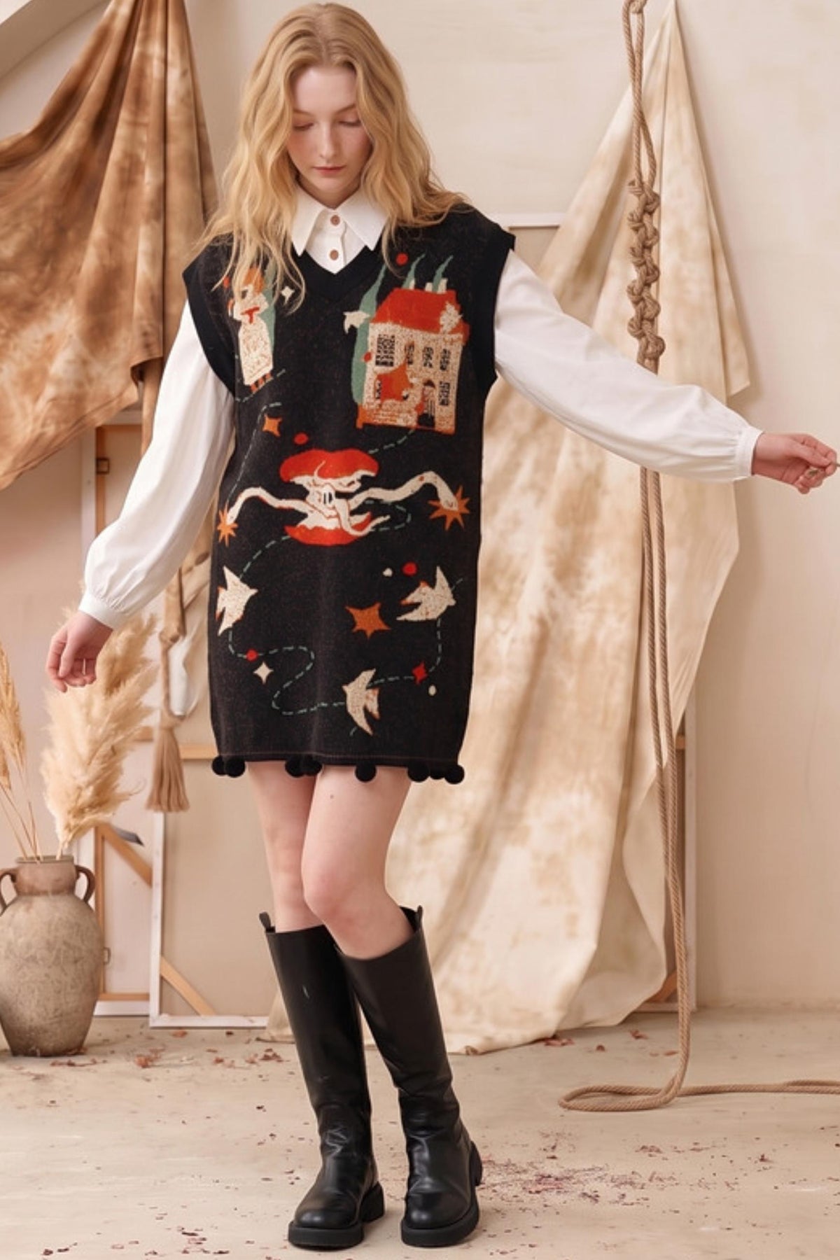 A woman modeling a black knit sweater vest with whimsical embroidered illustrations of a house, a skull and crossbones, stars, and birds, layered over a white collared shirt and black knee-high boots.