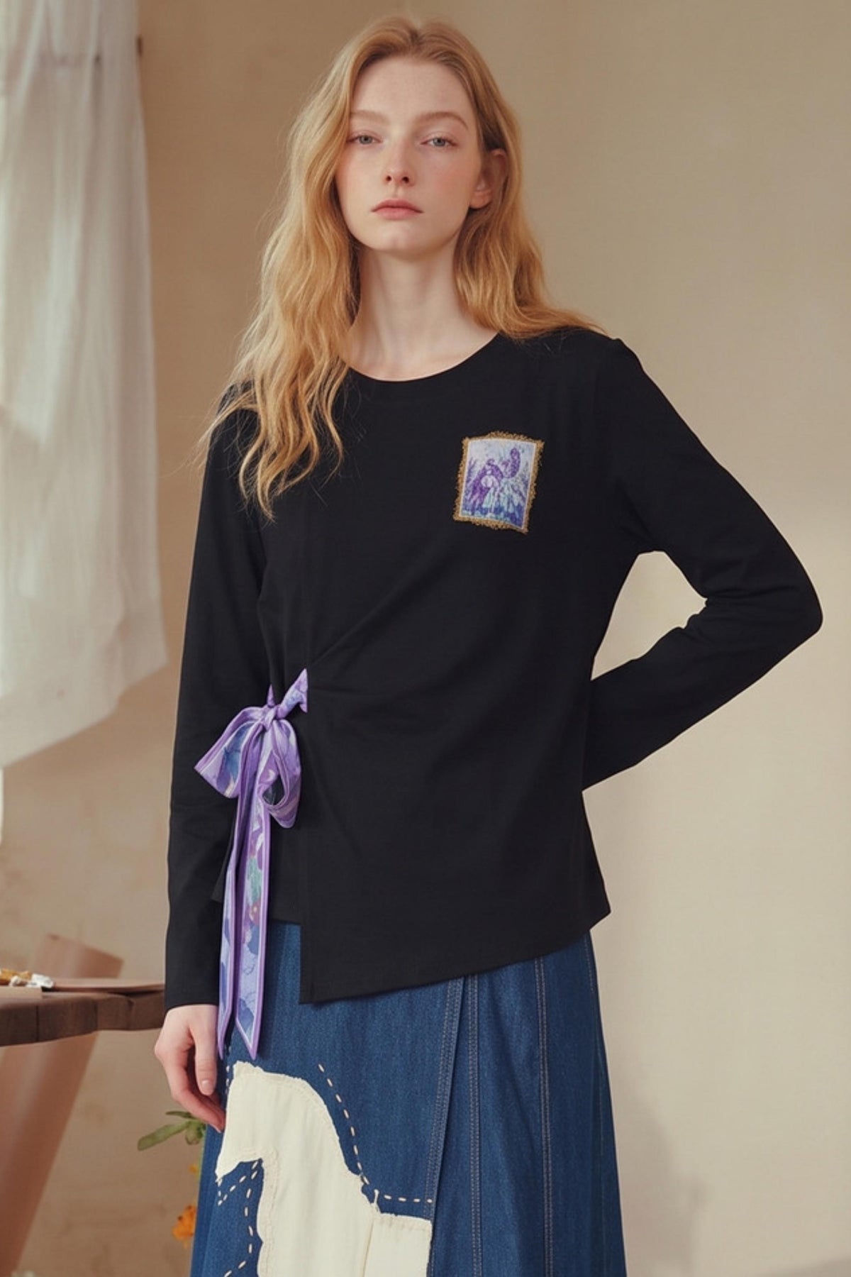 A woman modeling a black long-sleeve top with a small square graphic patch on the left chest and a purple patterned ribbon tied at the waist, paired with a blue and white patterned skirt in a softly lit indoor setting.
