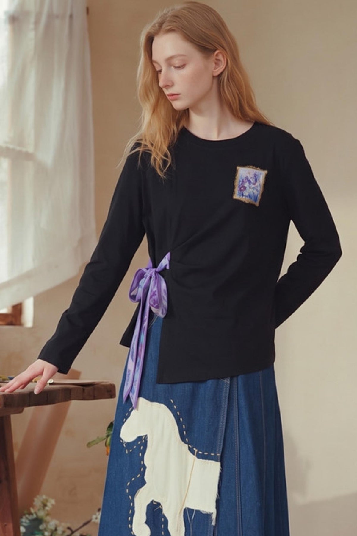 A woman wearing a black long-sleeve top with a purple ribbon tied at the waist and a blue skirt with a white horse graphic, standing indoors near a window.