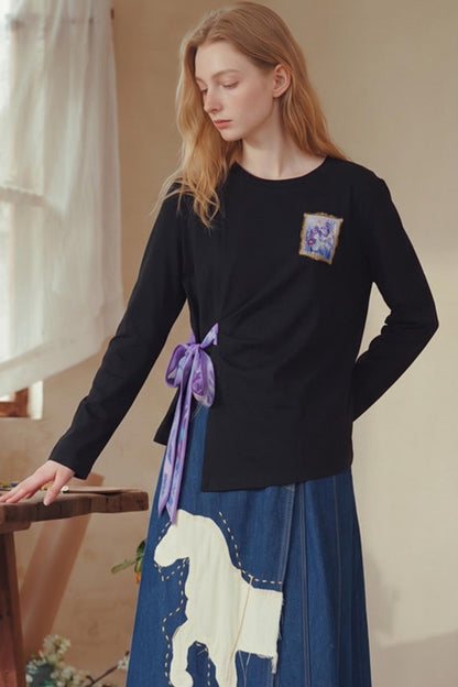 A woman wearing a black long-sleeve top with a purple ribbon tied at the waist and a blue skirt with a white horse graphic, standing indoors near a window.