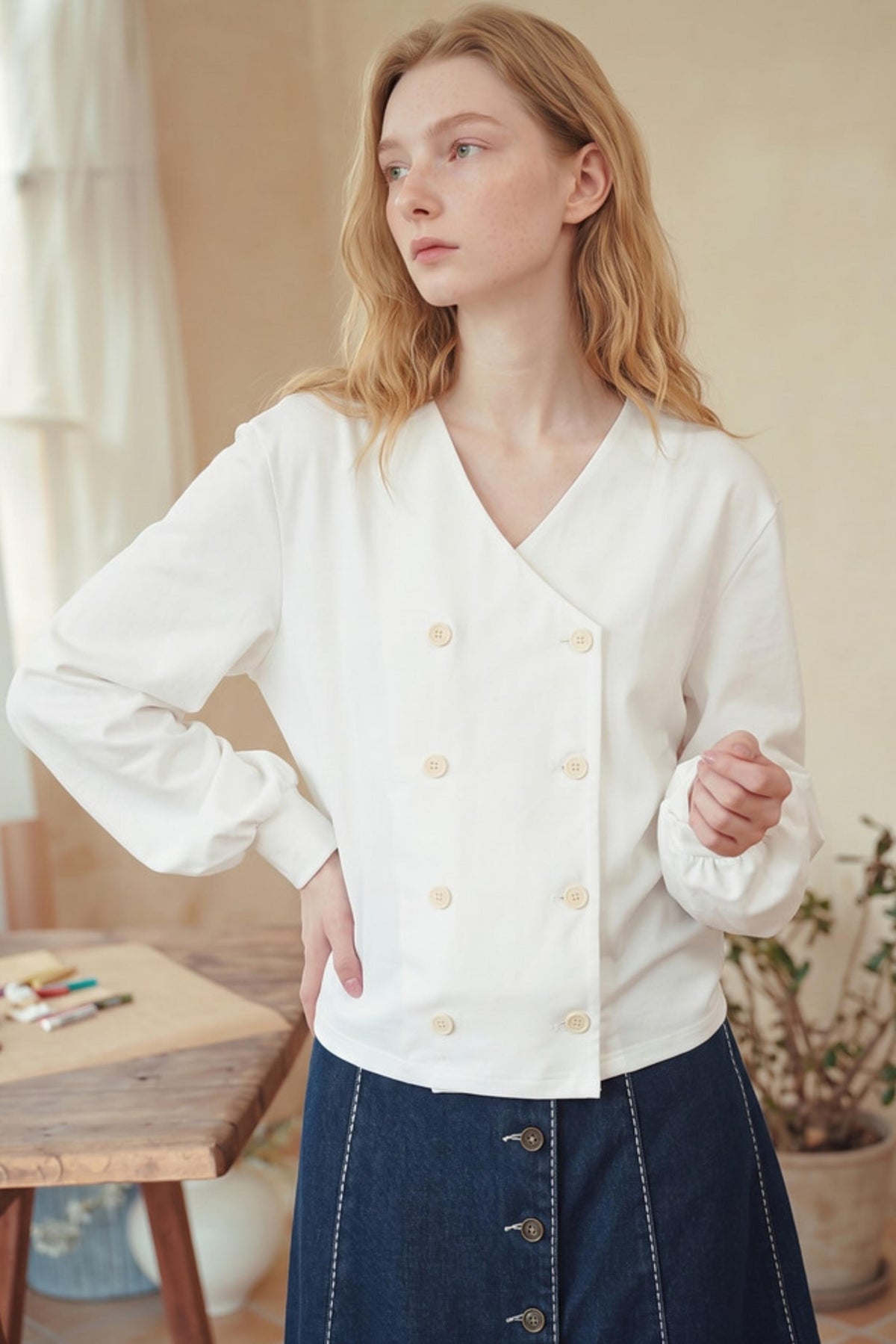 A woman wearing a white long-sleeve wrap-style blouse with decorative buttons, paired with a dark blue skirt, standing in a softly lit indoor setting.