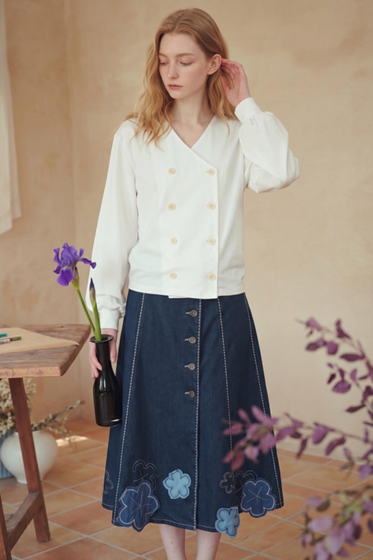 A woman stands indoors wearing a white long-sleeve wrap-style blouse with gold buttons and a dark blue midi skirt with floral embroidery and button details. She holds a small black vase with a purple iris in a warm, rustic room with beige walls and terracotta flooring.