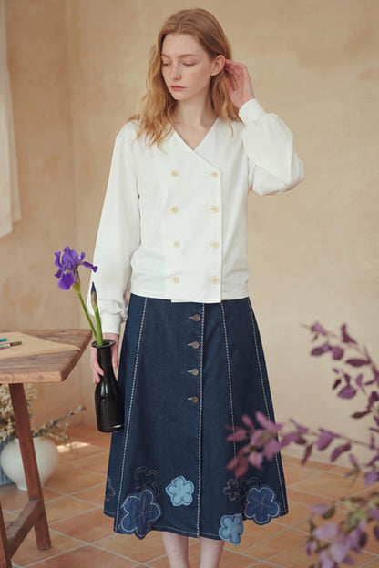 A woman stands indoors wearing a white long-sleeve wrap-style blouse with gold buttons and a dark blue midi skirt with floral embroidery and button details. She holds a small black vase with a purple iris in a warm, rustic room with beige walls and terracotta flooring.