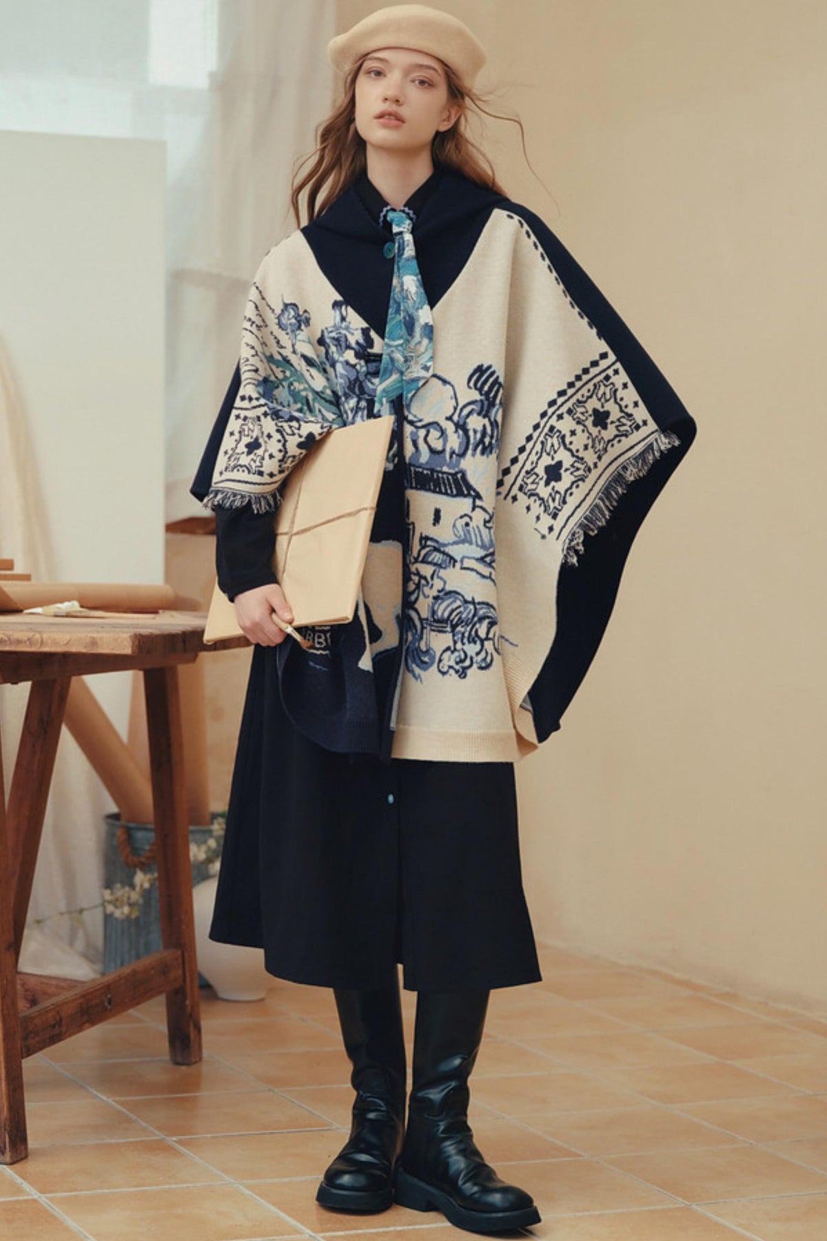 A woman stands indoors wearing a cream and navy patterned shawl over a black dress, with a beige beret, a blue patterned tie, a beige envelope-style clutch, and black knee-high boots.