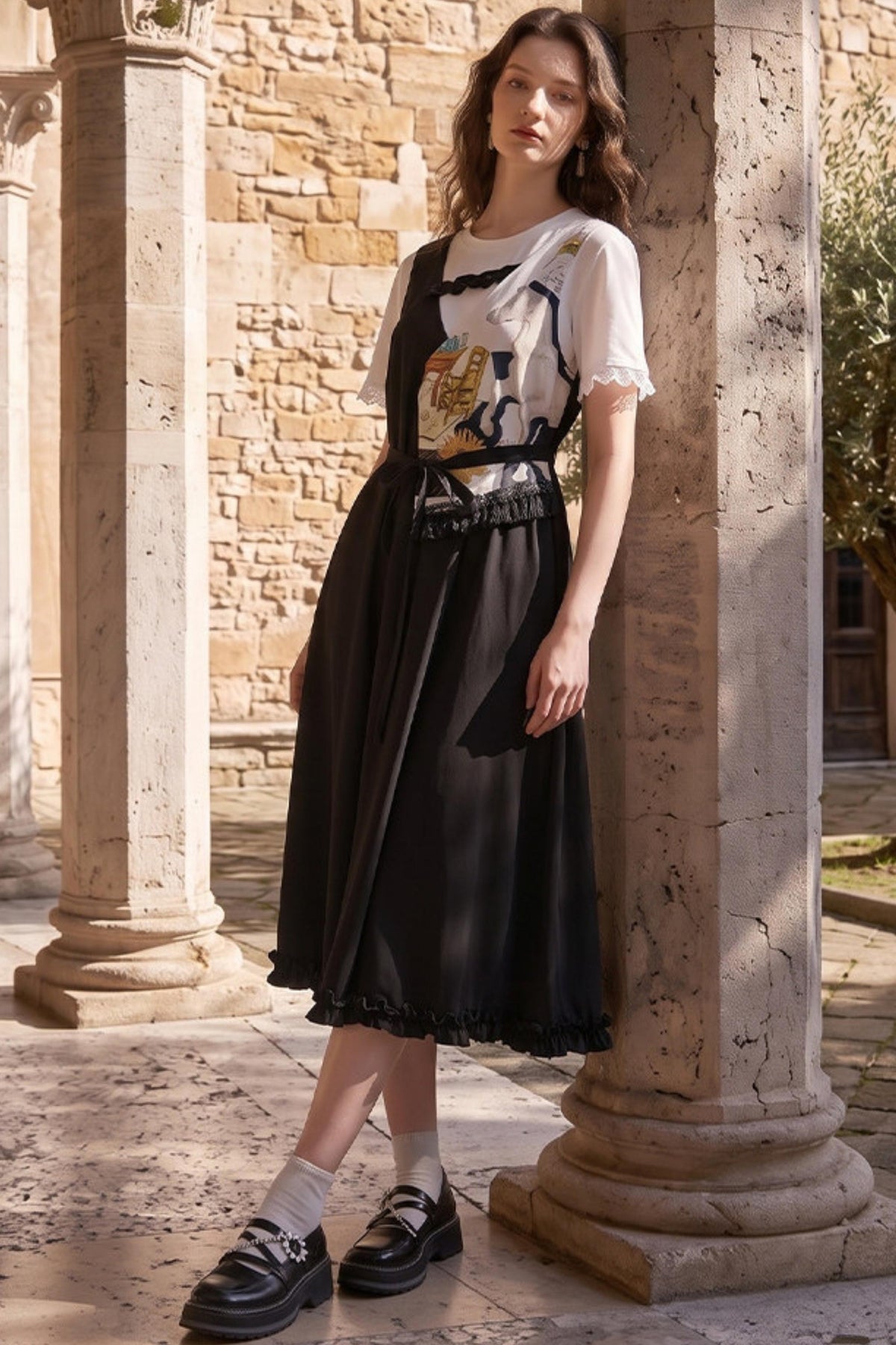 A woman stands in a sunlit stone courtyard between large columns, wearing a black midi dress with a white short-sleeve top layered underneath and black loafers.