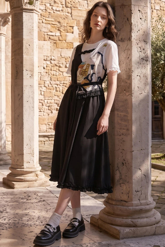 A woman stands in a sunlit stone courtyard between large columns, wearing a black midi dress with a white short-sleeve top layered underneath and black loafers.