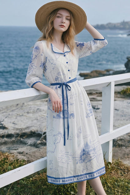A woman wearing a long white midi dress with a blue architectural print, blue trim, and a matching blue tie belt, standing on a white railing with the ocean in the background.