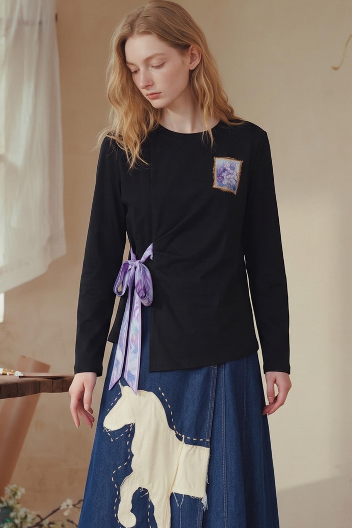 A woman wearing a black long-sleeve top with a purple embroidered patch and a blue skirt featuring a cream-colored horse patch, standing in a softly lit indoor setting.