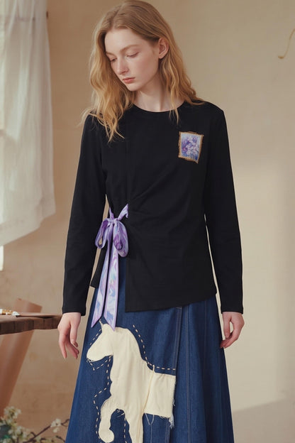A woman wearing a black long-sleeve top with a purple embroidered patch and a blue skirt featuring a cream-colored horse patch, standing in a softly lit indoor setting.