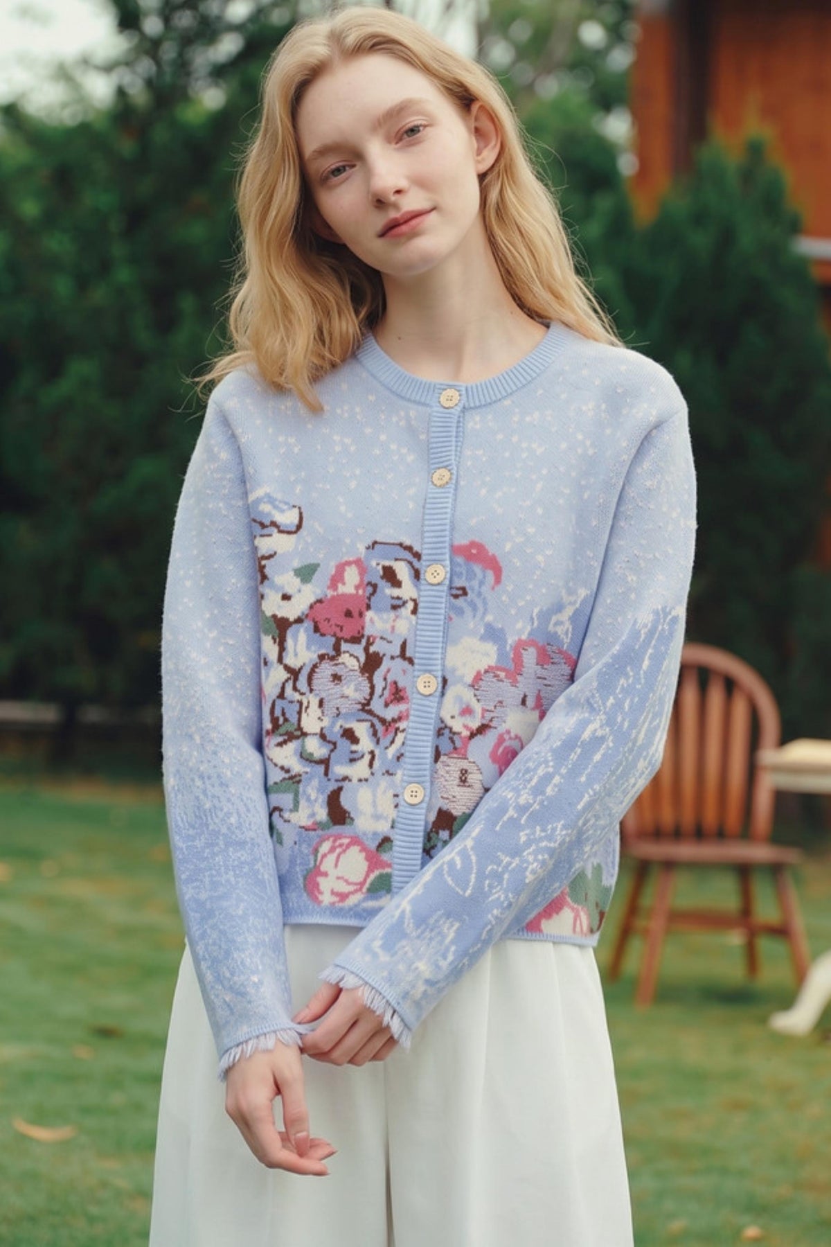 A woman wearing a light blue knit cardigan with a colorful floral and animal print on the front, standing outdoors in a grassy area with trees and a wooden chair in the background.