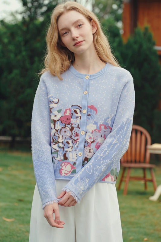 A woman wearing a light blue knit cardigan with a colorful floral and animal print on the front, standing outdoors in a grassy area with trees and a wooden chair in the background.