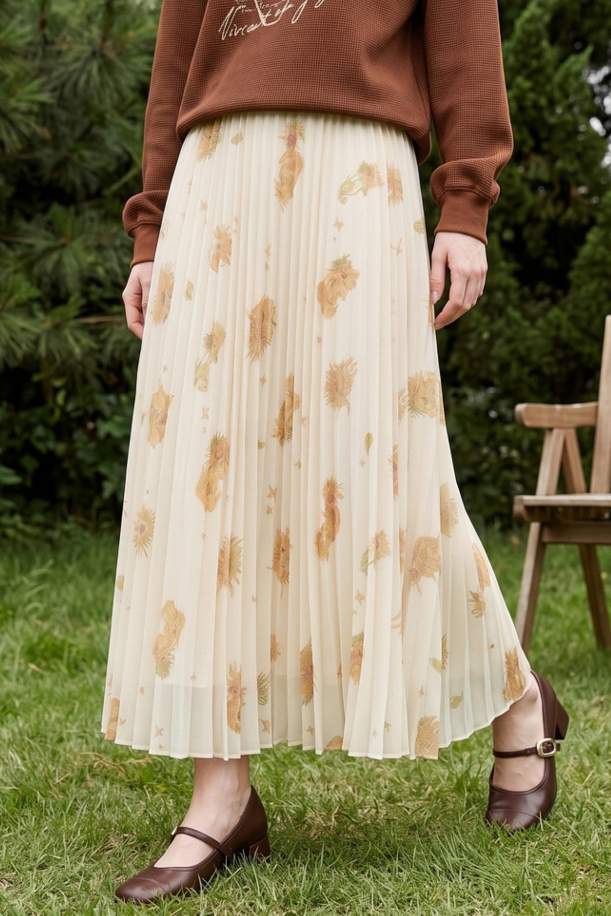A person standing outdoors on grass wearing a long pleated midi skirt in cream with a tan floral print, a brown sweatshirt, and brown Mary Jane flats. Trees and a wooden chair are visible in the background.