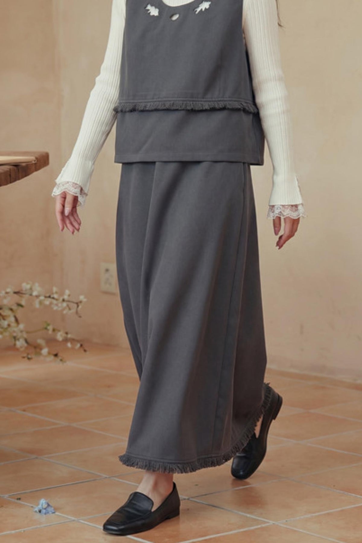 A person wearing a dark gray sleeveless vest and matching wide-leg skirt set with fringe, layered over a cream ribbed long-sleeve top with lace cuffs, paired with black loafers in a warm beige indoor setting.