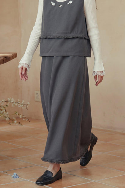 A person wearing a dark gray sleeveless vest and matching wide-leg skirt set with fringe, layered over a cream ribbed long-sleeve top with lace cuffs, paired with black loafers in a warm beige indoor setting.