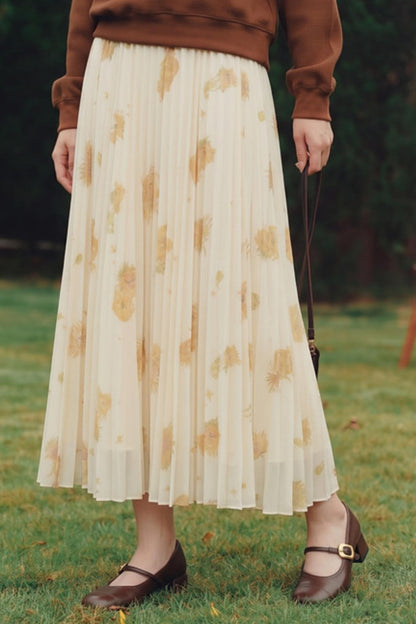 A person standing outdoors on grass wearing a cream pleated midi skirt with a tan leaf print, a brown sweater, and brown Mary Jane shoes.