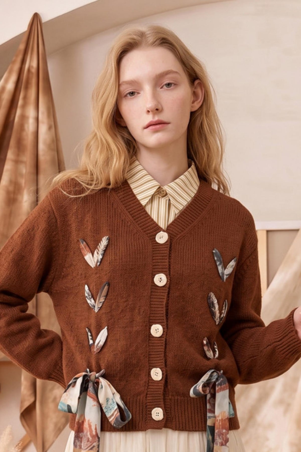 A woman wearing a brown ribbed cardigan with feather and ribbon embellishments over a striped collared shirt, posed indoors against a warm neutral backdrop.