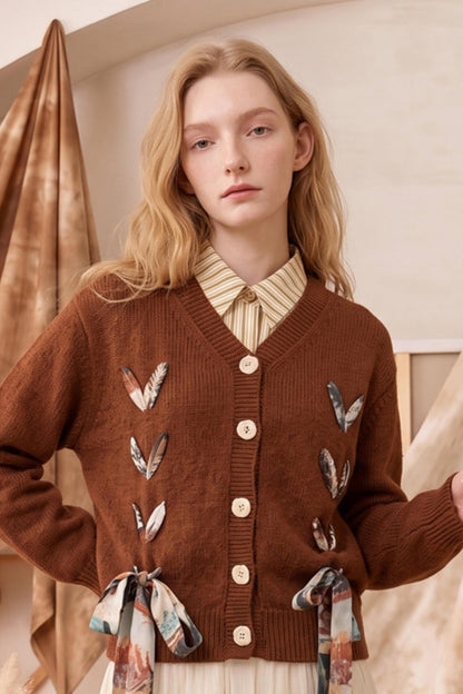 A woman wearing a brown ribbed cardigan with feather and ribbon embellishments over a striped collared shirt, posed indoors against a warm neutral backdrop.