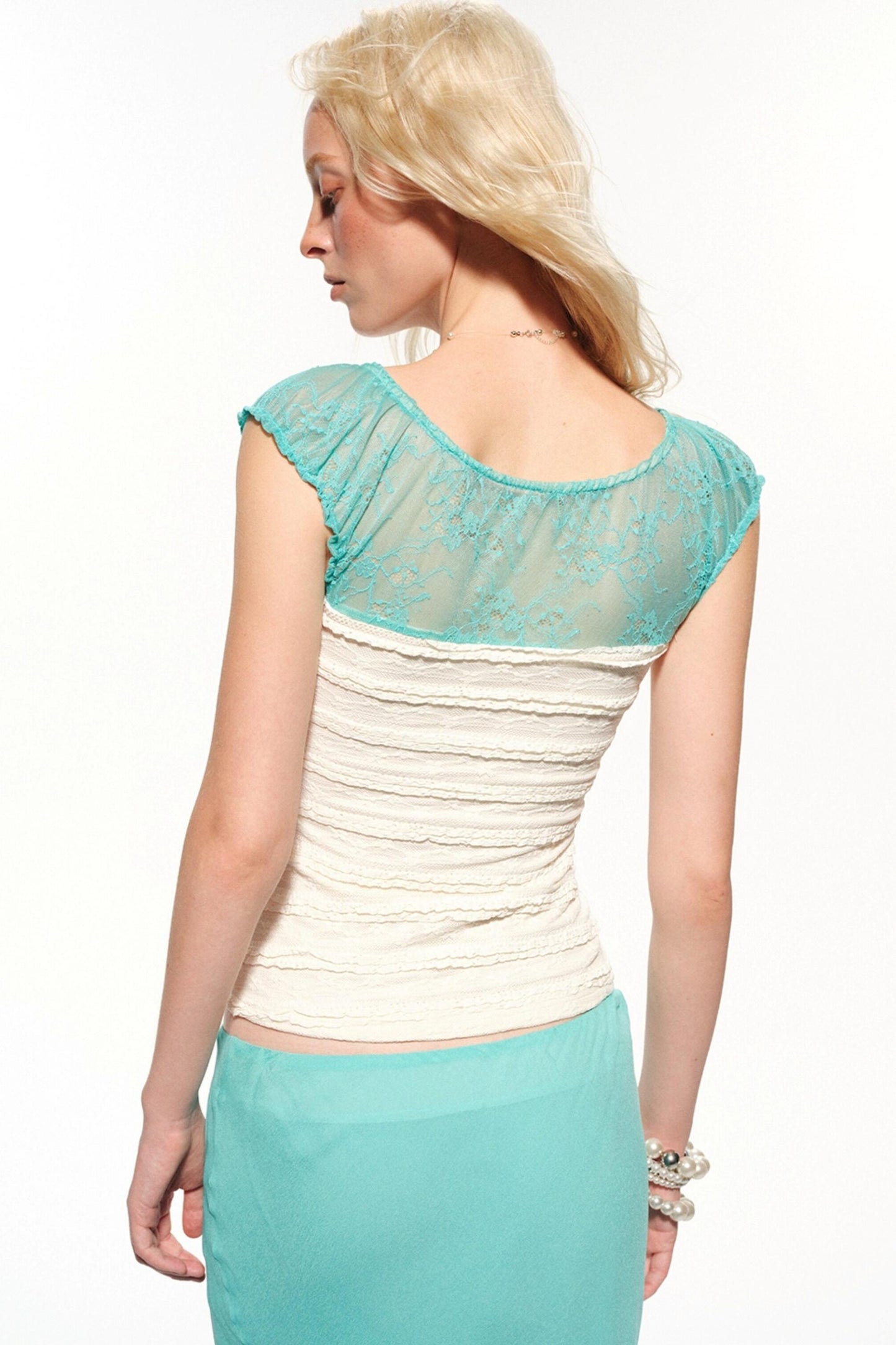 A blonde model shown from the back wearing a two-tone top with a turquoise lace cap-sleeve yoke and an off-white ruched body, paired with a matching turquoise skirt, against a plain white background.