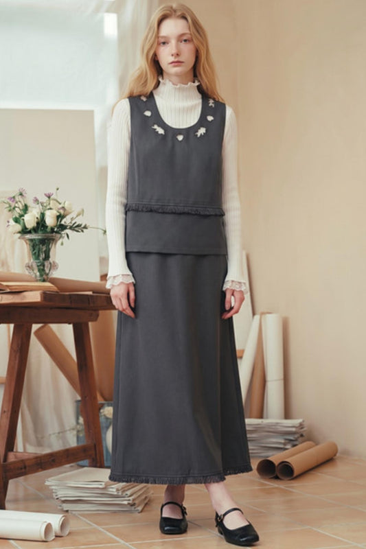 A woman stands indoors wearing a dark charcoal gray sleeveless vest over a white long-sleeve ruffled turtleneck, paired with a matching long skirt and black Mary Jane flats. The setting is a warm, softly lit room with a wooden table, flowers, and rolled papers in the background.