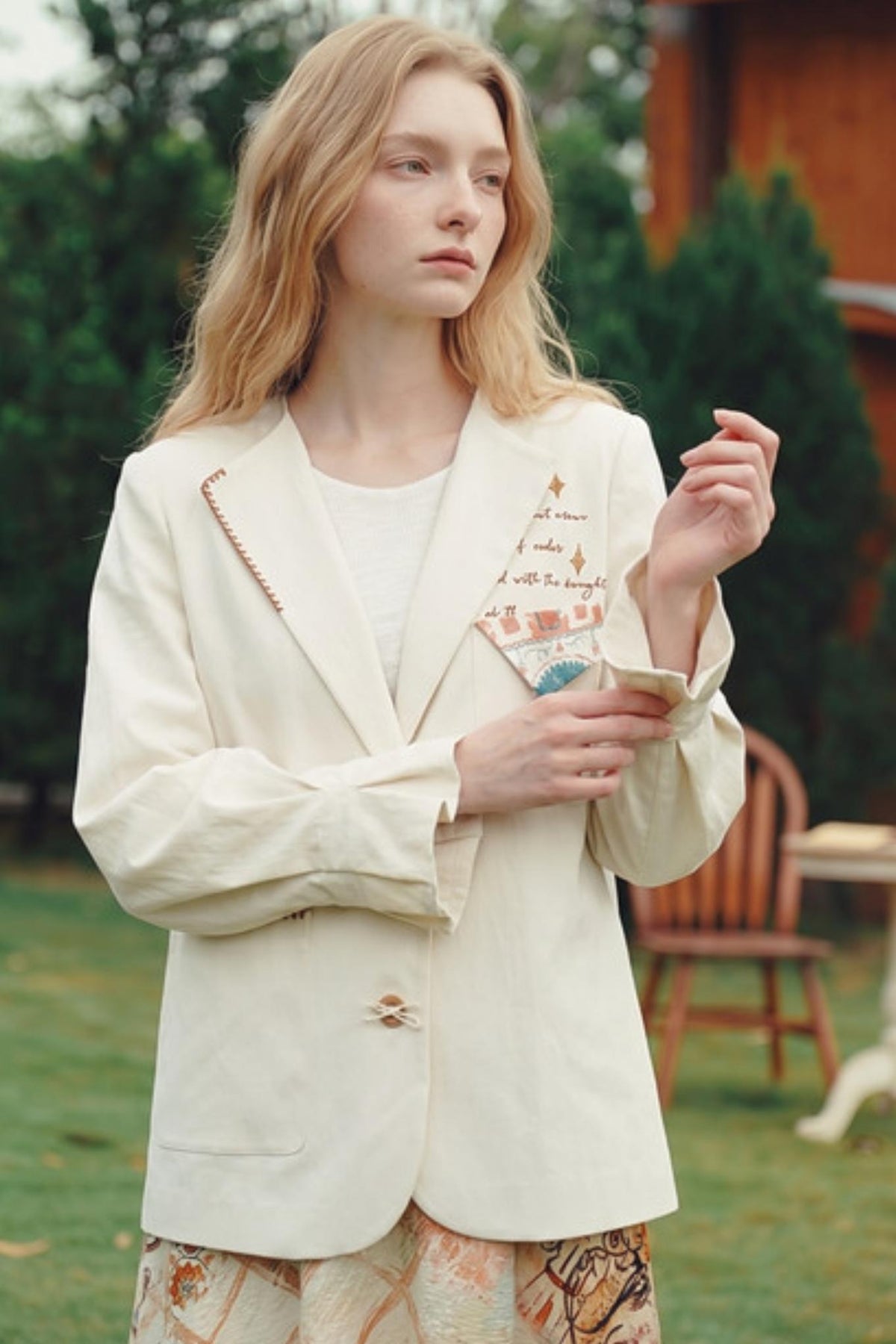 A woman standing outdoors in a garden wearing a cream-colored blazer with decorative embroidery and a single button closure, paired with a white top and patterned skirt.