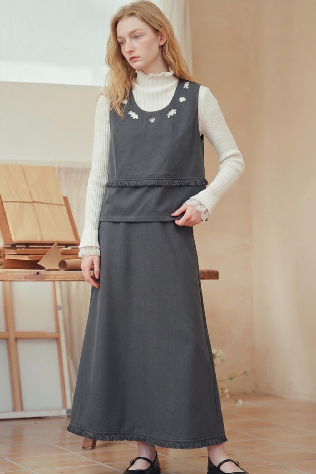 A woman models a dark gray sleeveless pinafore-style dress layered over a white ribbed turtleneck sweater. The dress features small white embroidered details around the neckline and a ruffled waist seam.