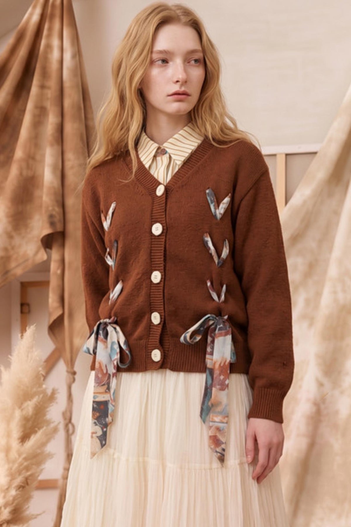 A woman wearing a brown cardigan with decorative bird motifs and matching patterned tie details at the waist, styled over a cream striped shirt and pleated cream skirt in a warm neutral indoor setting.