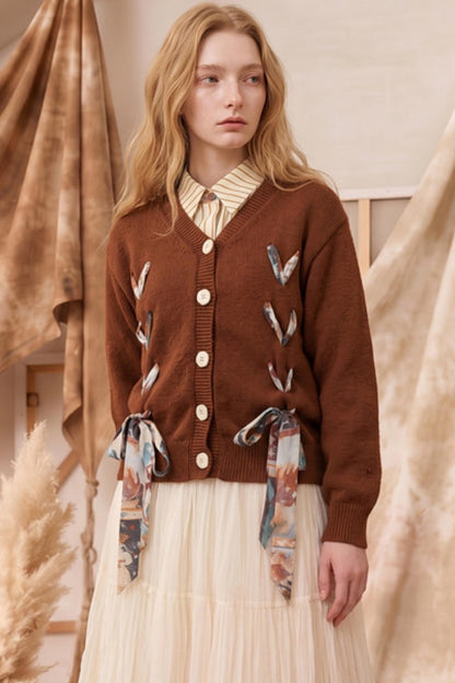 A woman wearing a brown cardigan with decorative bird motifs and matching patterned tie details at the waist, styled over a cream striped shirt and pleated cream skirt in a warm neutral indoor setting.