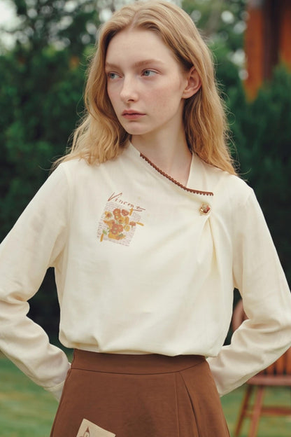 A young woman with blonde hair wearing a cream-colored long-sleeve blouse with a floral graphic and a brown pleated skirt, standing outdoors.
