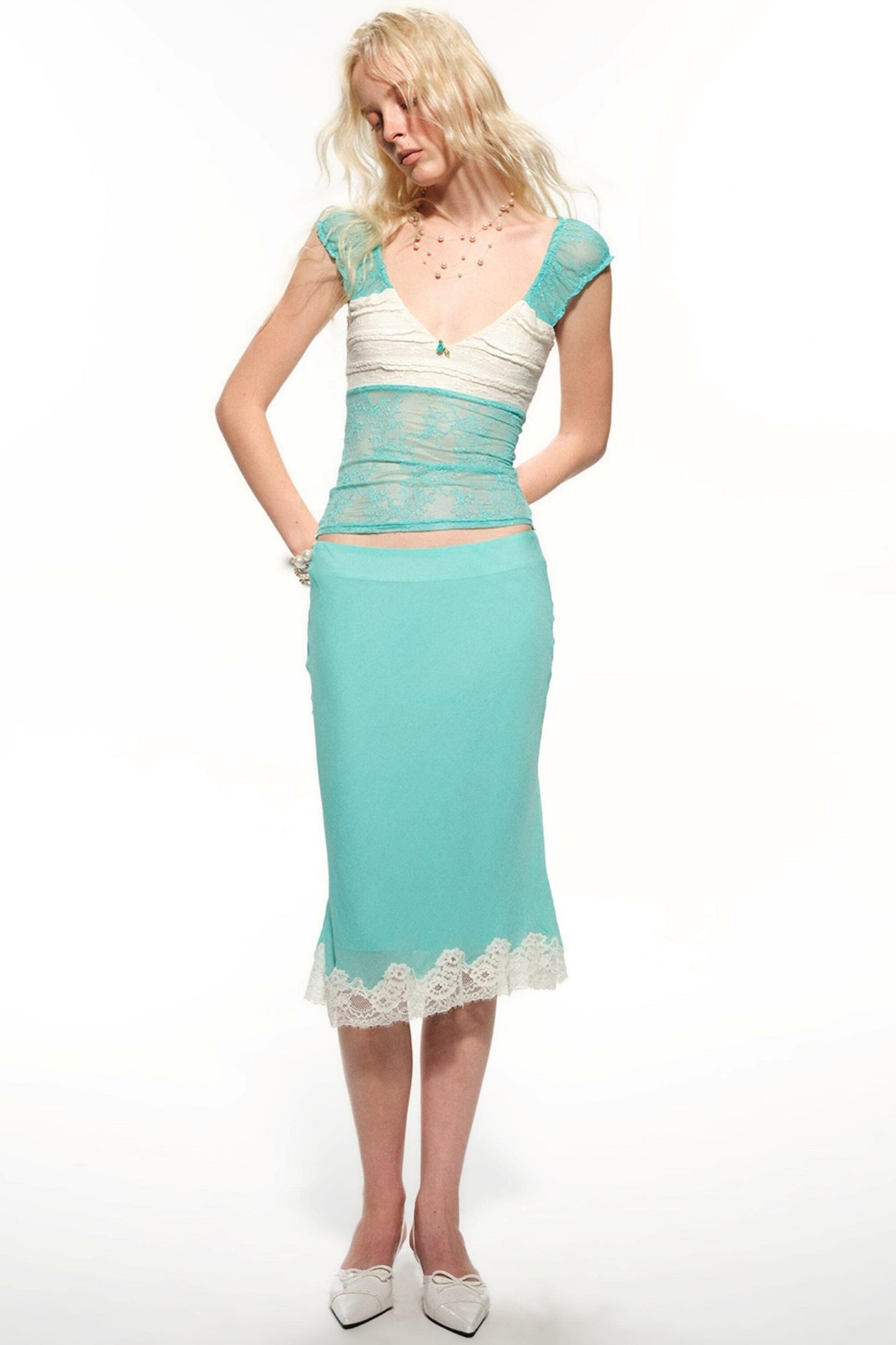 A woman modeling a two-piece turquoise and white outfit consisting of a lace-trimmed crop top and matching midi skirt, against a plain white studio background.