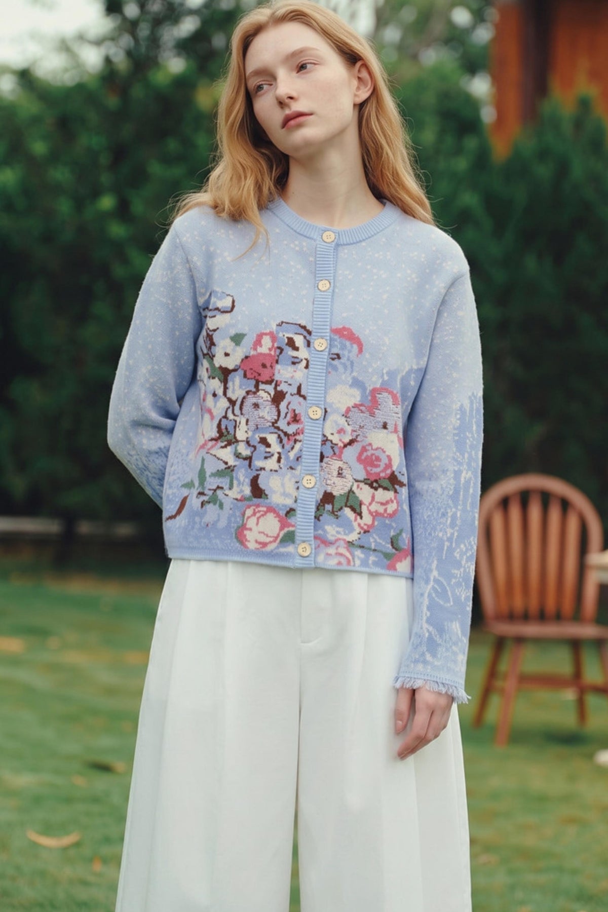 A woman standing outdoors in a garden wearing a light blue cardigan with a large floral and bird pattern on the front, paired with white wide-leg pants.