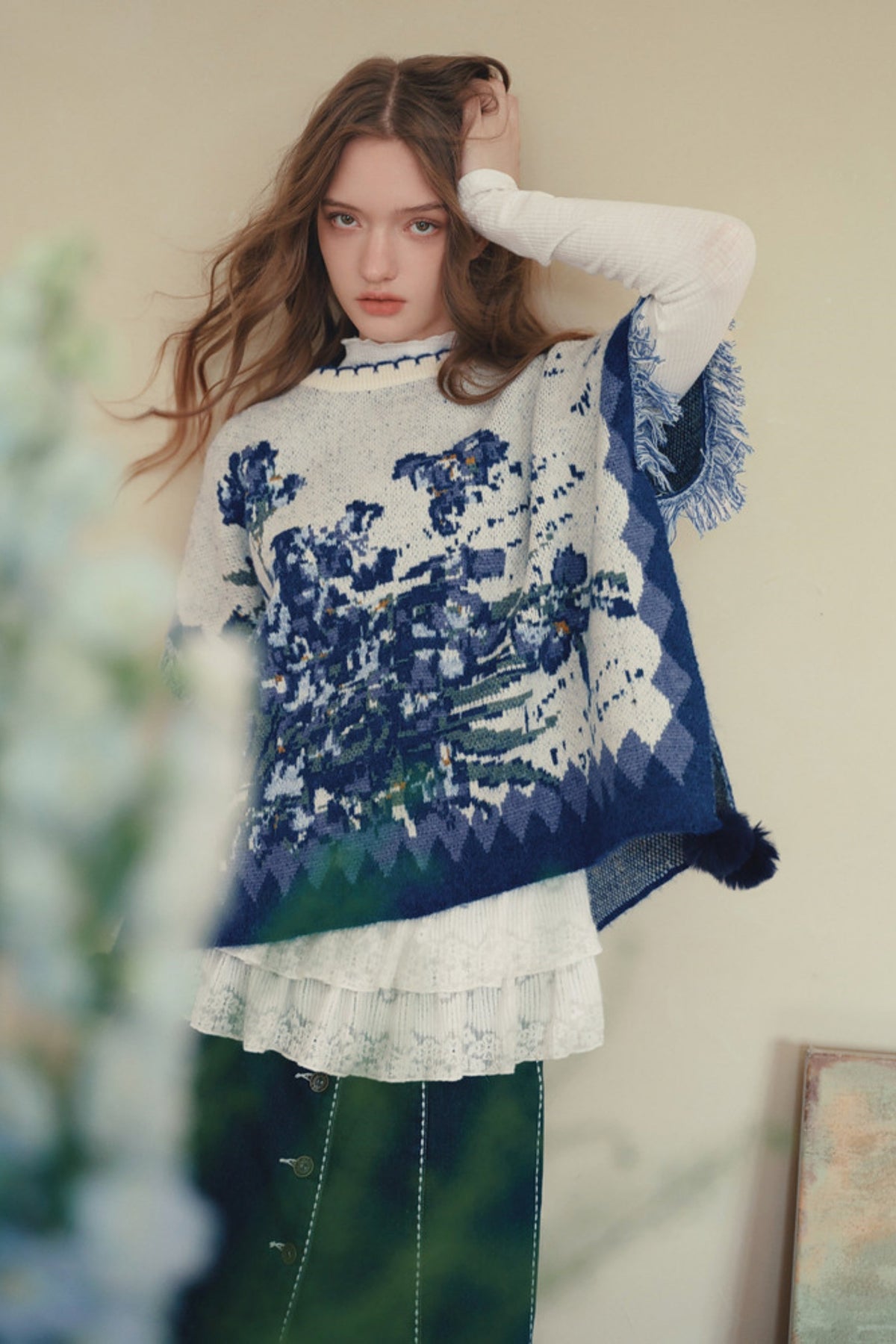 A woman wearing a blue and white patterned knit poncho over a white long-sleeve top and a green skirt, posing in front of a neutral beige wall.