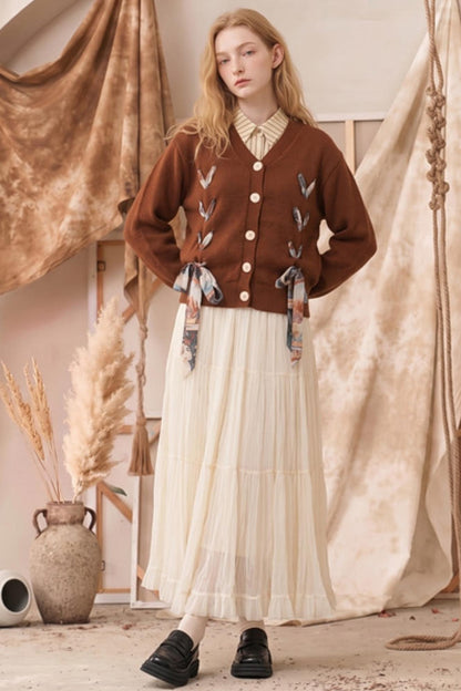 A woman stands in a styled studio setting wearing a brown button-front cardigan with embroidered leaf details and patterned ribbon ties, paired with a long cream pleated skirt and black loafers.