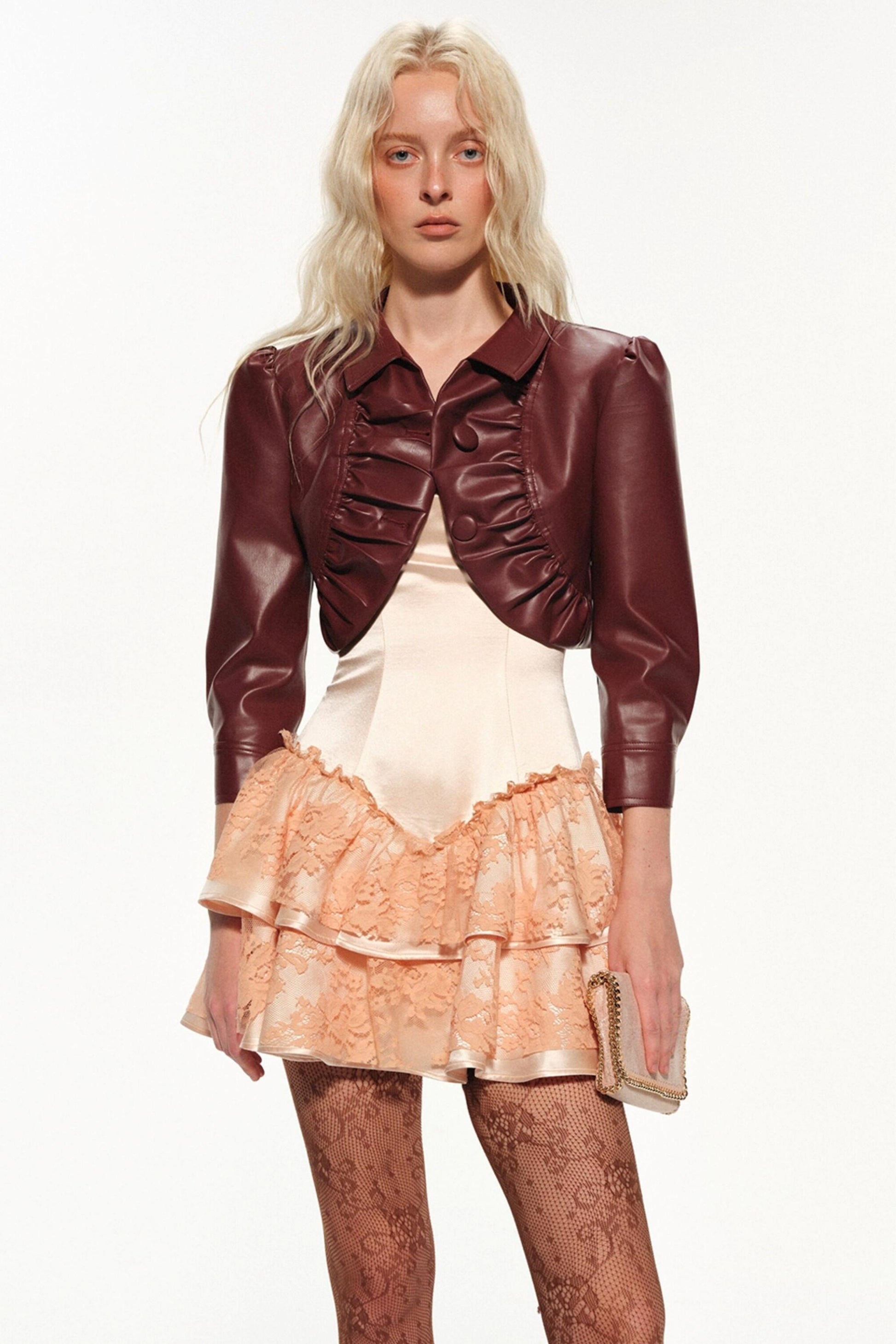 A blonde model wearing a dark burgundy faux-leather cropped jacket with ruffle details over a cream satin top and a peach lace ruffled mini skirt, holding a small metallic clutch against a plain white background.