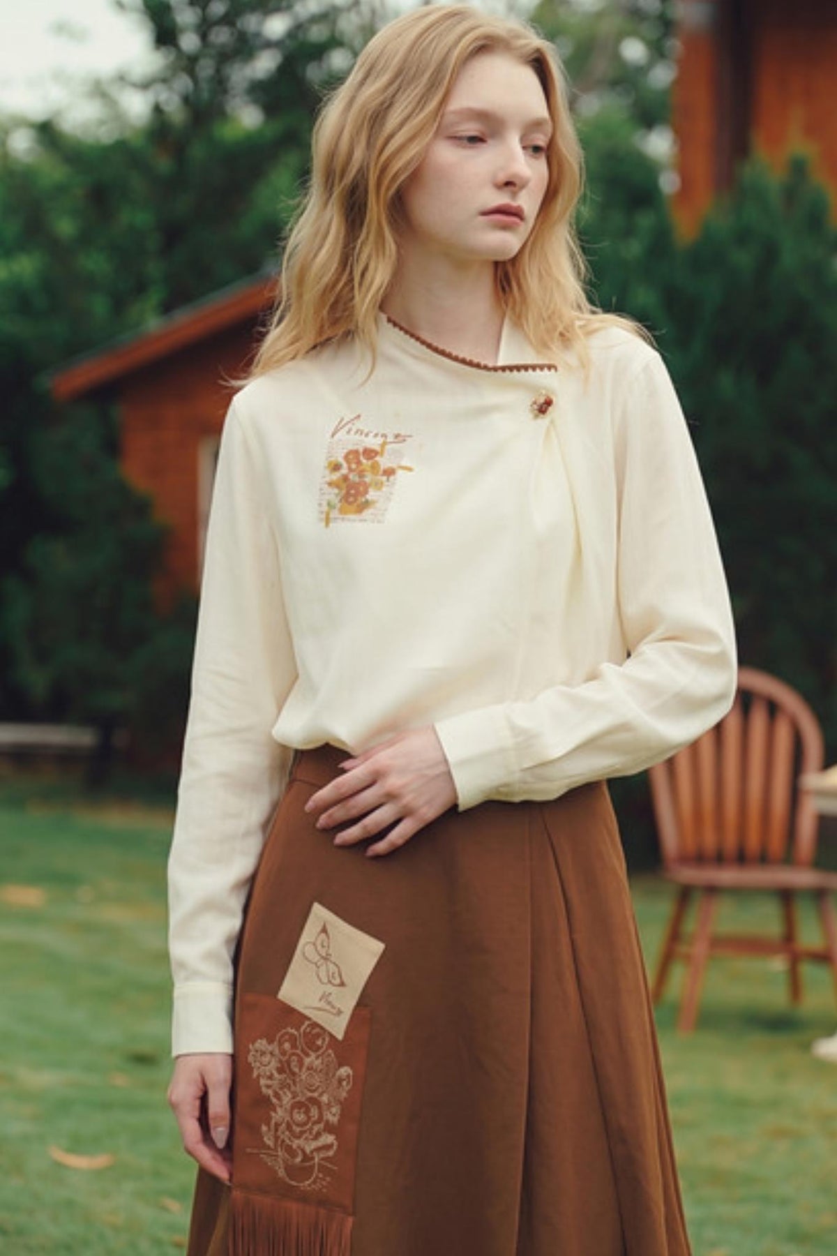 A woman standing outdoors in a garden wearing a cream long-sleeve blouse with a small embroidered chest design and a brown skirt with fringe and embroidery.