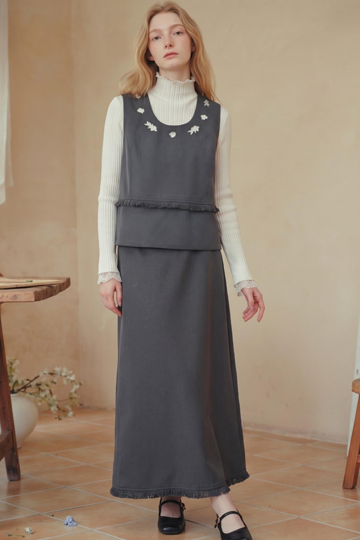 A woman stands indoors wearing a dark gray sleeveless layered dress over a white ribbed turtleneck, with black Mary Jane shoes. The room has a warm beige wall and terracotta tile floor.